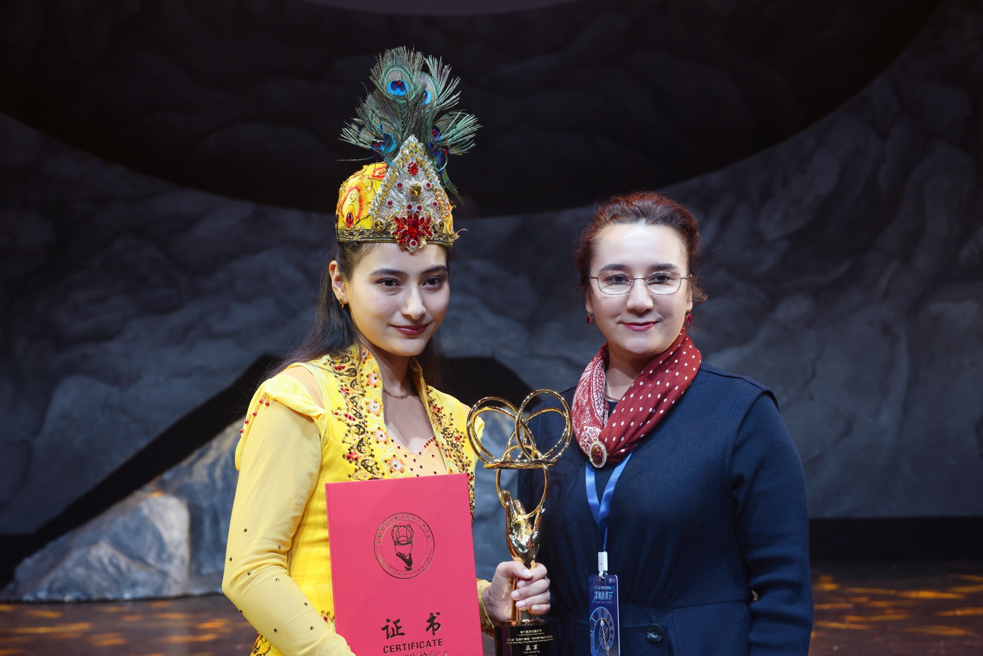 第六届深圳魔术节颁奖台上,古扎丽阿衣手持证书与「金魔环」奖杯,与师父茹仙古丽合影