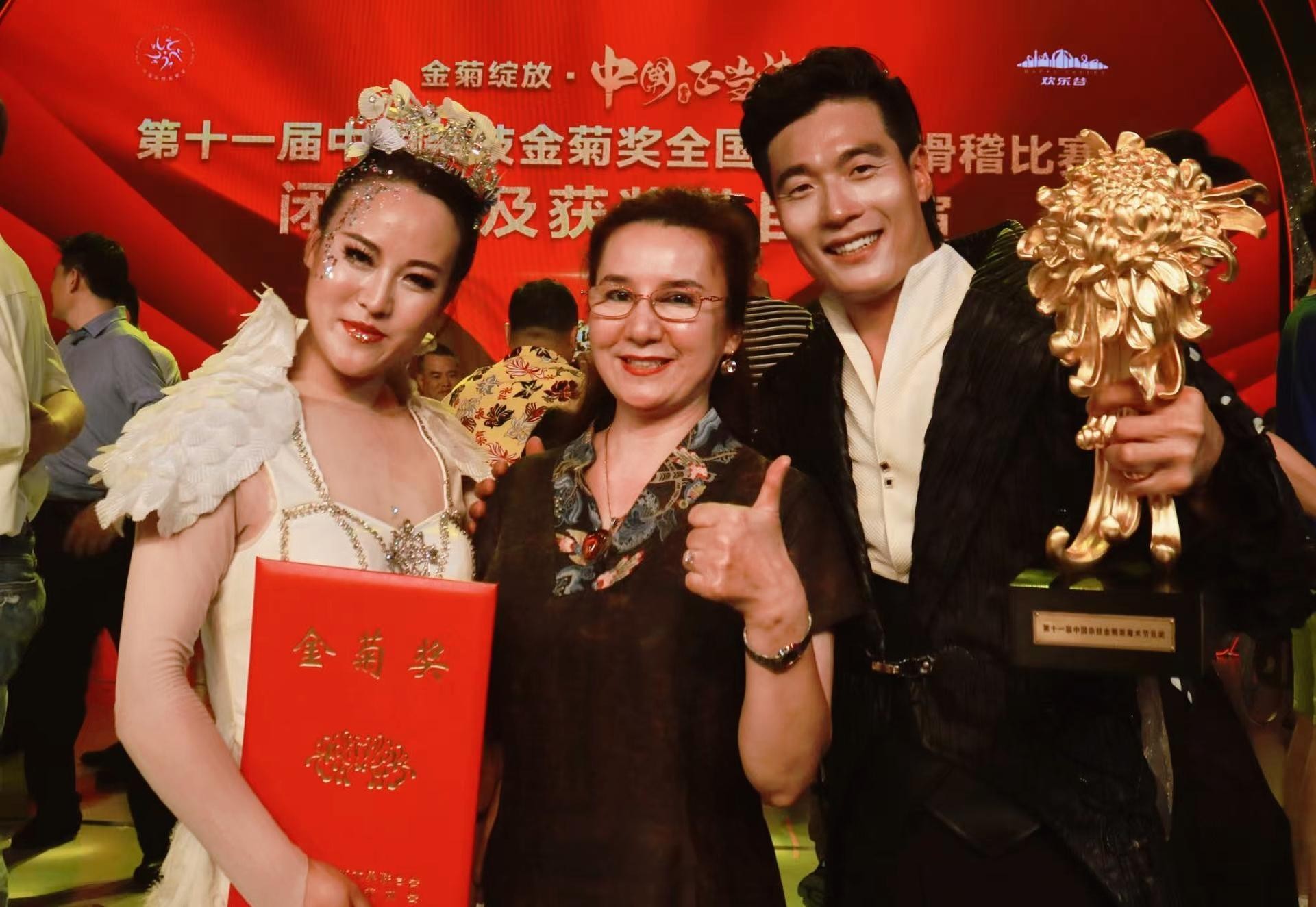 第十一届中国杂技金菊奖闭幕式,茹仙古丽居中,右侧胡玲珑手捧金菊花奖杯,左侧一位身着白色羽毛演出服的女士手持金菊奖证书
