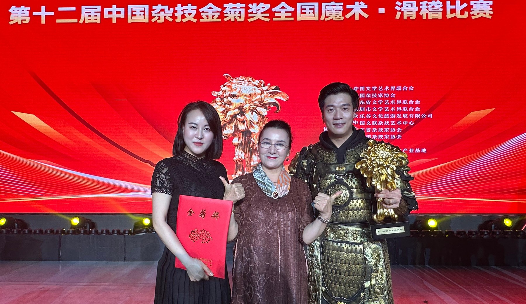 第十二届中国杂技金菊奖颁奖现场,茹仙古丽居中,右侧胡玲珑着金甲手捧金菊奖杯,左侧一位女士手持金菊奖证书