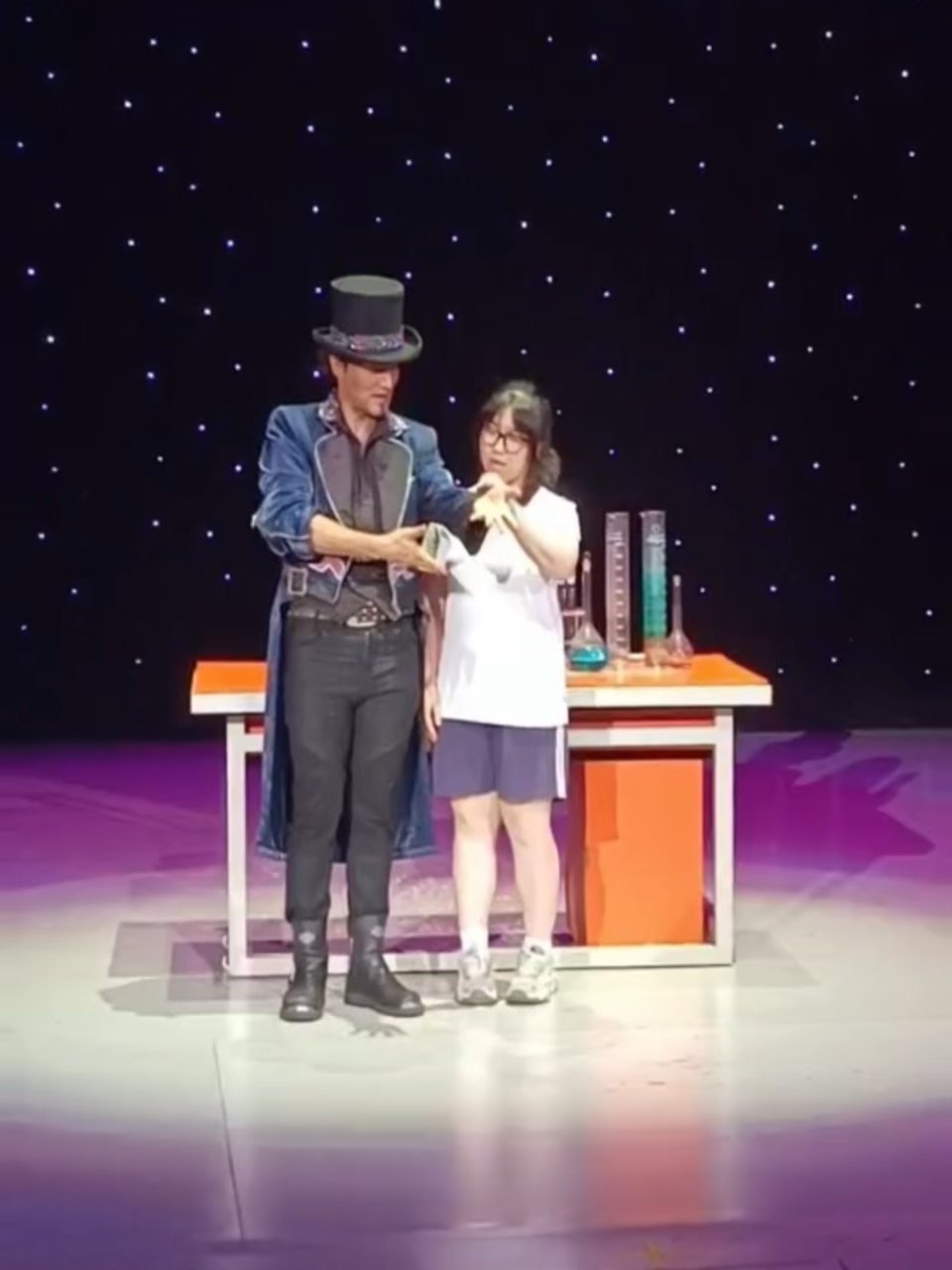 舞台上他与一名女童并立于摆着道具的小桌前